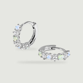 A pair of silver hoops shows alternating iridescent and clear stones against a soft gray surface.