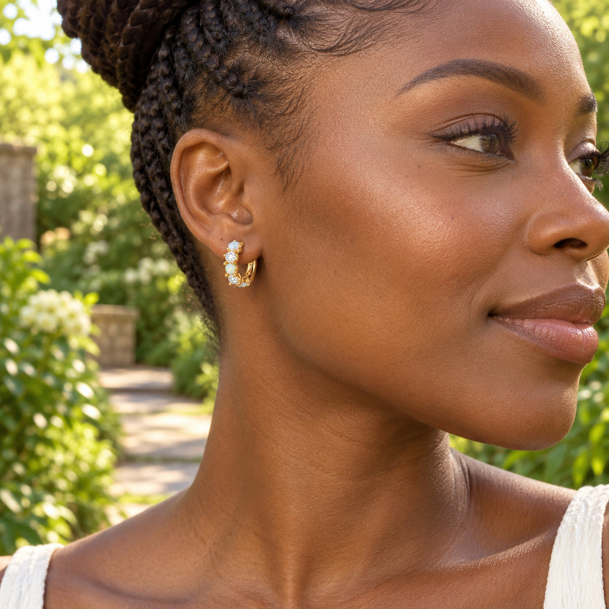 A yellow gold hoop with blue, green, and clear stones is worn beside a garden path.