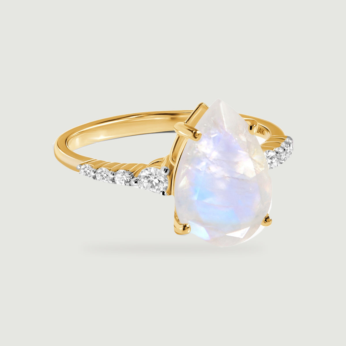 A yellow gold ring with a pear-shaped moonstone and diamond shoulders rests on a light grey surface.