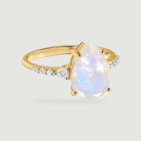 A yellow gold ring with a pear-shaped moonstone and diamond shoulders rests on a light grey surface.