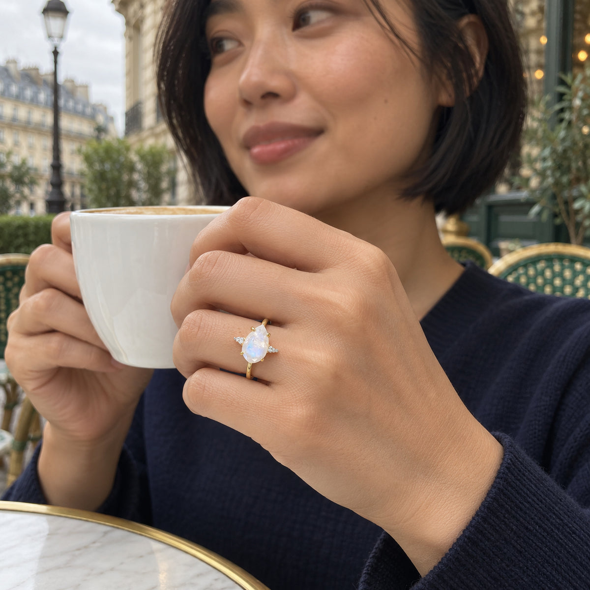 A woman wears a yellow gold moonstone and diamond ring while holding a white coffee cup at an outdoor cafe.