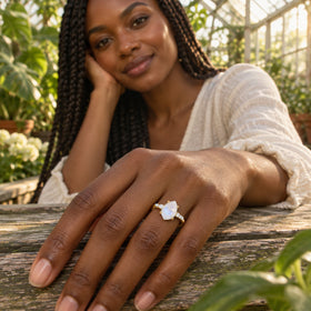 A yellow gold ring with a pear-shaped moonstone is worn on a hand resting on a wooden table in a greenhouse.
