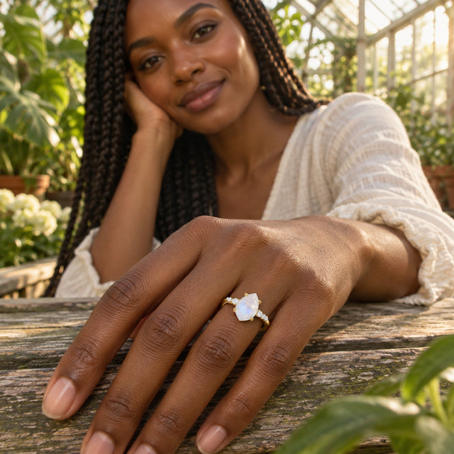 A yellow gold ring with a pear-shaped moonstone is worn on a hand resting on a wooden table in a greenhouse.