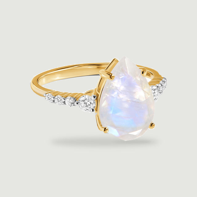 A pear-shaped moonstone ring in yellow gold sits above a soft shadow on a light gray surface.