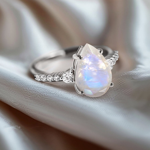 A white gold moonstone ring rests on folded pale gray fabric with the pear-shaped stone facing forward.