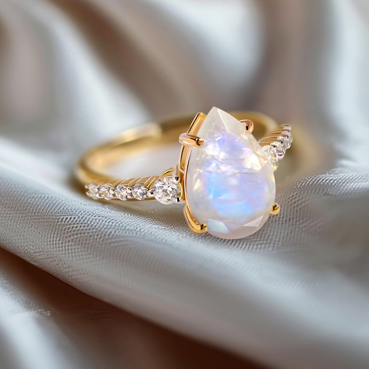 A yellow gold moonstone ring rests on folded pale gray fabric with the pear-shaped stone facing forward.