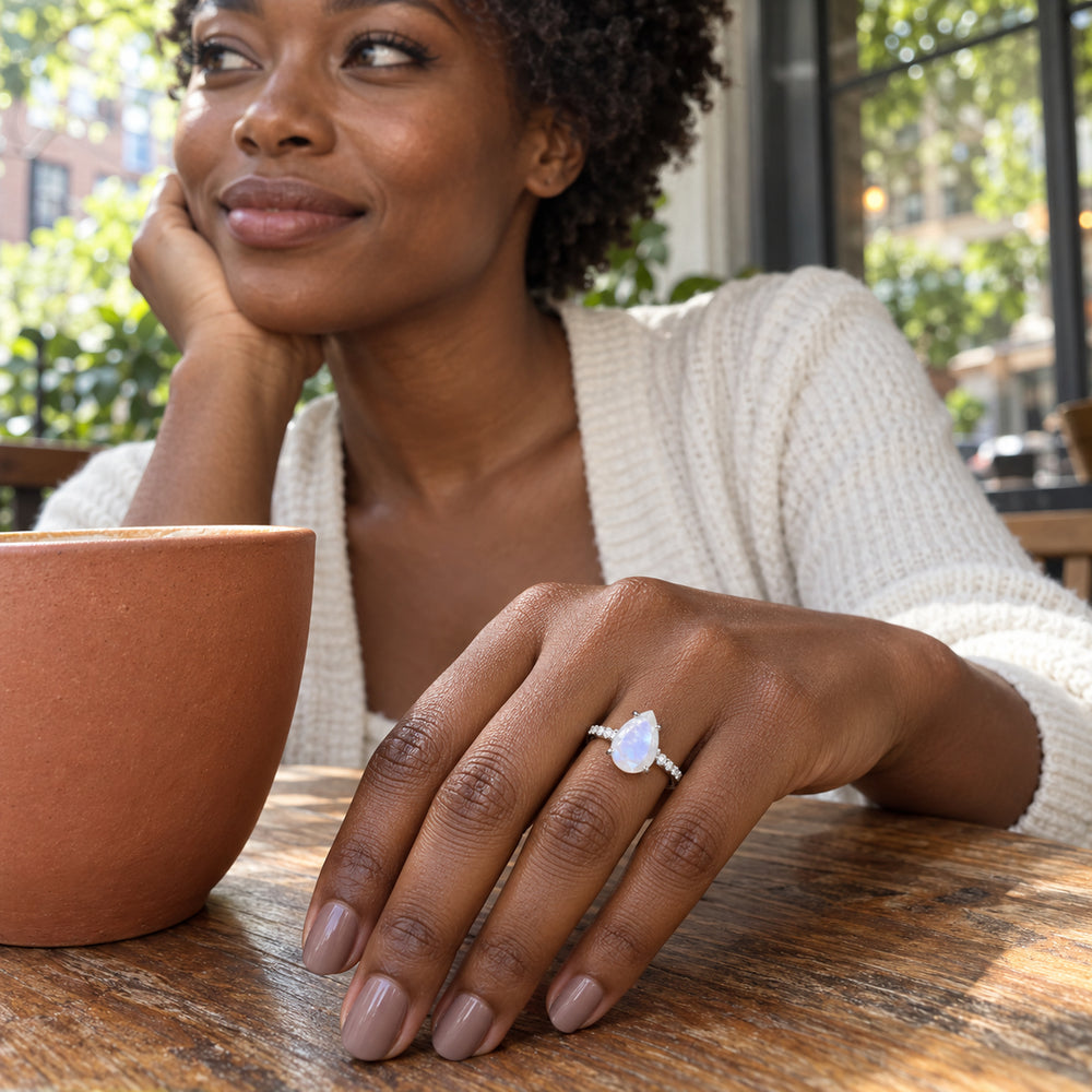 A pear-shaped moonstone ring in white gold is worn on a hand beside a terracotta cup at a cafe table.