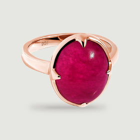 An oval deep-pink cabochon ring with a polished rose-gold band rests on a light gray surface.