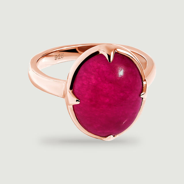 An oval deep-pink cabochon ring with a polished rose-gold band rests on a light gray surface.
