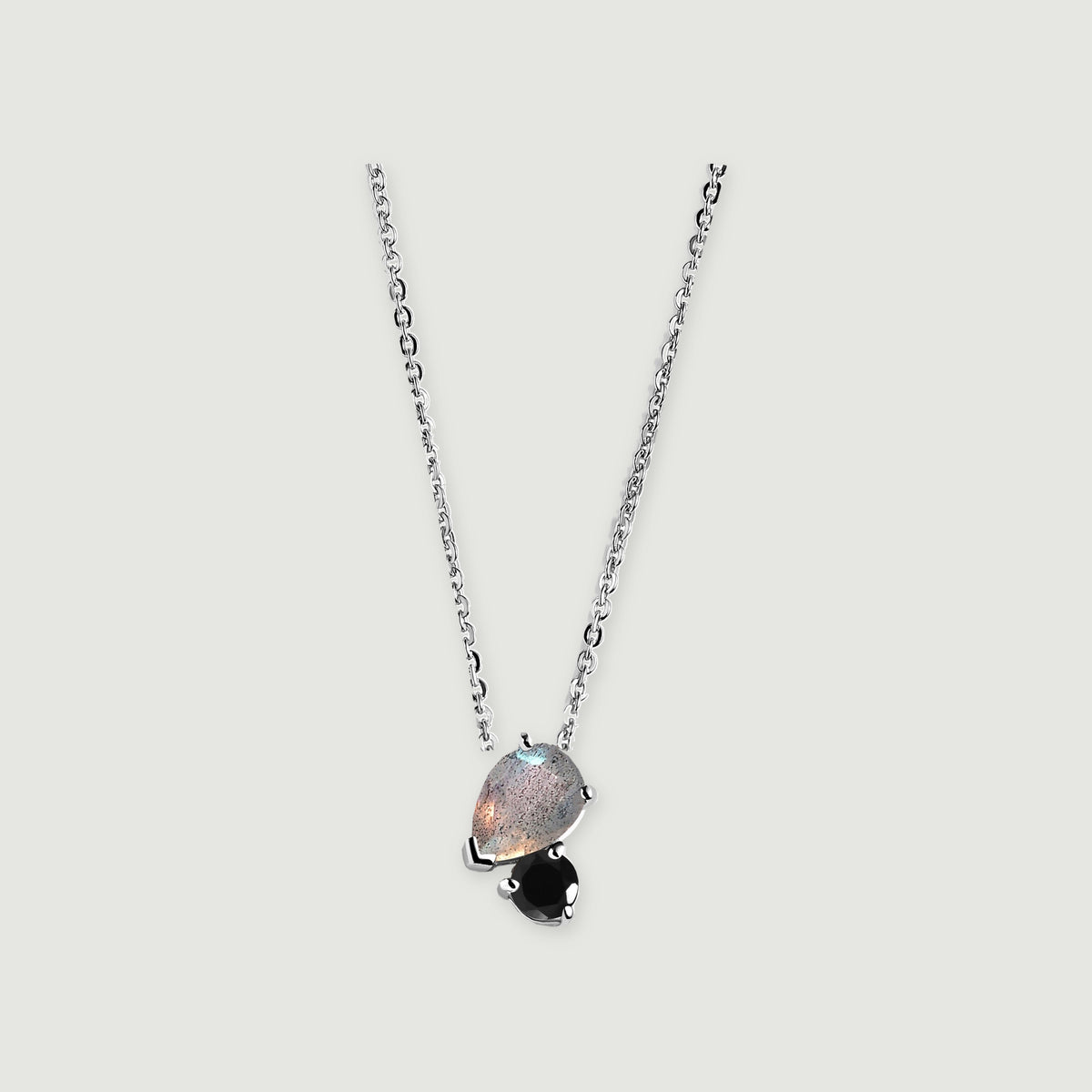 A silver chain holds a pear-shaped gray labradorite beside a round black obsidian stone on a light gray background.