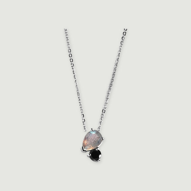 A silver chain holds a pear-shaped gray labradorite beside a round black obsidian stone on a light gray background.