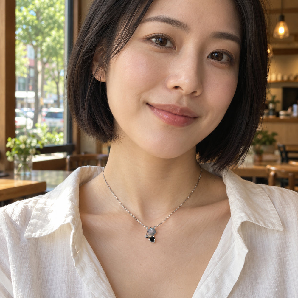 A woman with a short dark bob wears the silver two-stone necklace with a white blouse beside a cafe window.