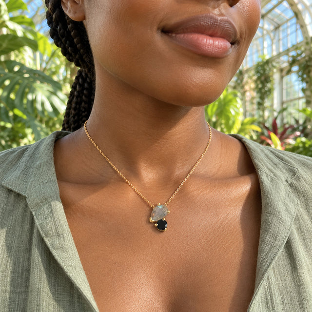 A yellow gold necklace with gray labradorite and black obsidian rests above a sage shirt amid sunlit conservatory foliage.