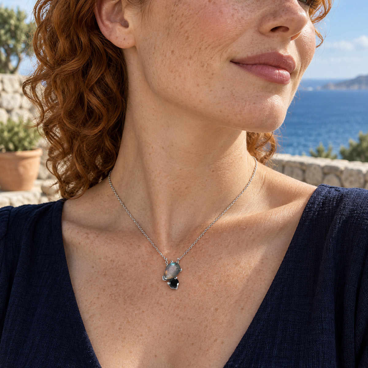 A silver pear-shaped labradorite and round black obsidian pendant rests on a freckled neckline above a navy top on a seaside terrace.