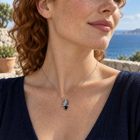 A silver pear-shaped labradorite and round black obsidian pendant rests on a freckled neckline above a navy top on a seaside terrace.