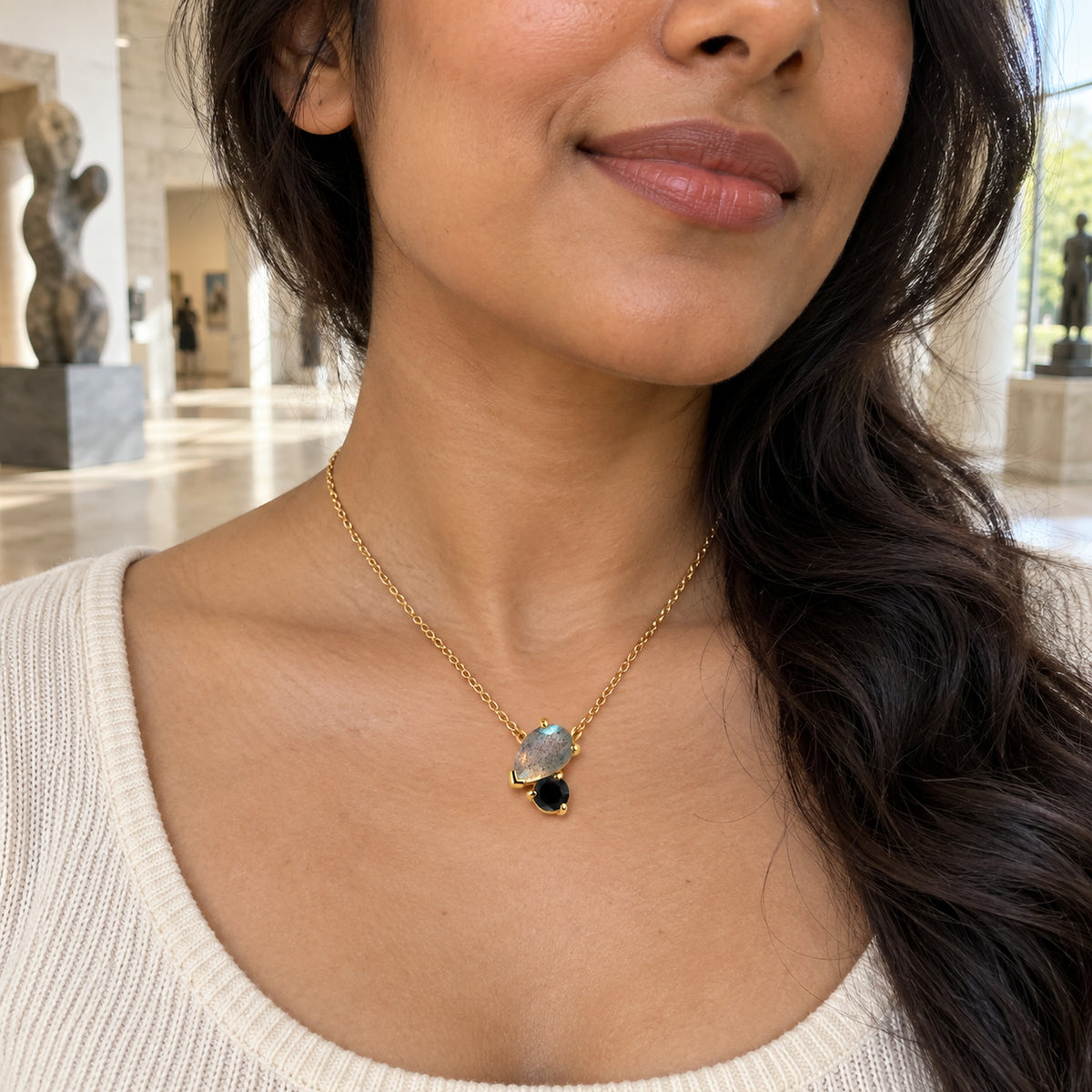 The yellow gold two-stone pendant is worn above a cream knit neckline with blurred sculptures behind the model.
