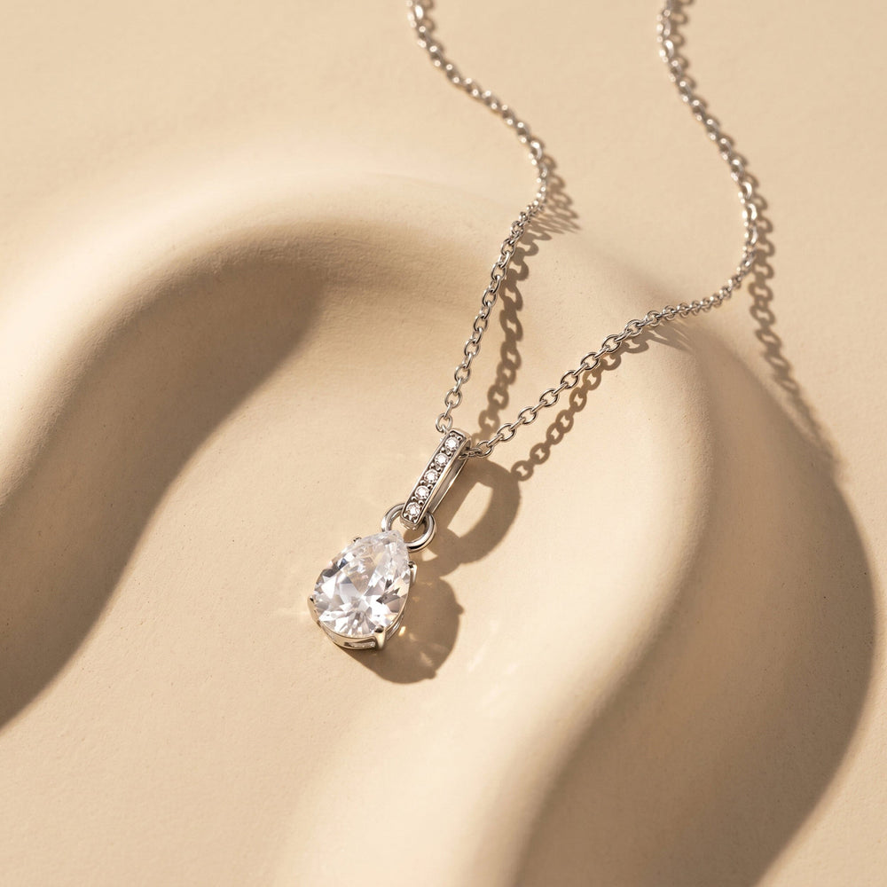 A silver chain and clear teardrop pendant lie across a textured cream surface with curved shadows.
