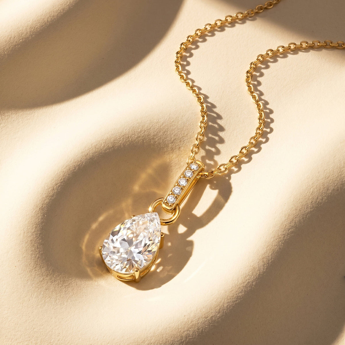 A gold chain curves across a cream surface above a pear-shaped pendant with a row of small round stones.