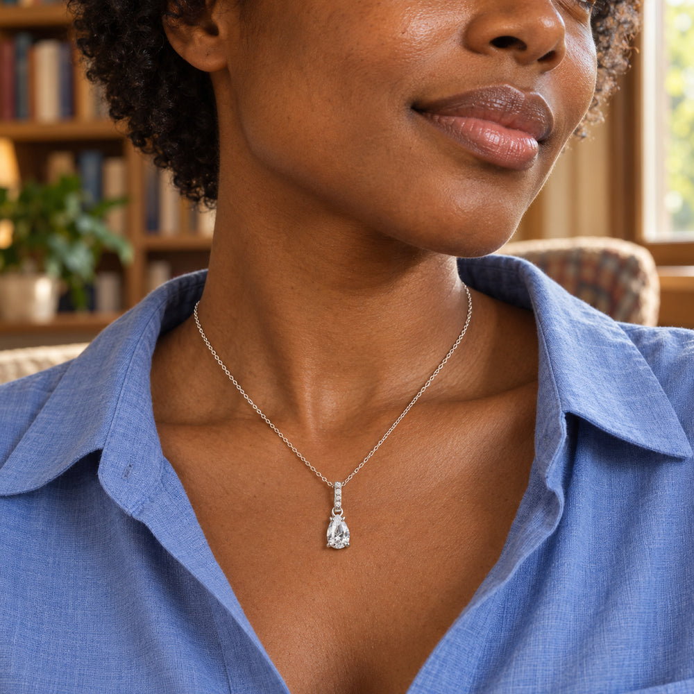 A woman in a blue open-collar shirt wears the silver pear-shaped pendant beside a window with bookshelves behind her.