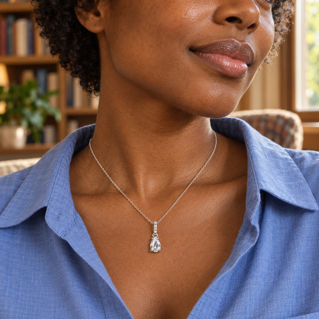 A woman in a blue open-collar shirt wears the silver pear-shaped pendant beside a window with bookshelves behind her.