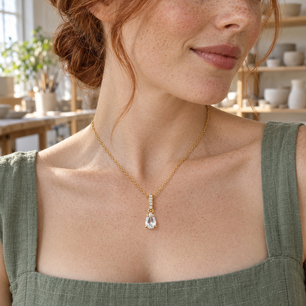 A red-haired woman in a green linen dress wears the gold teardrop necklace in a pottery studio.