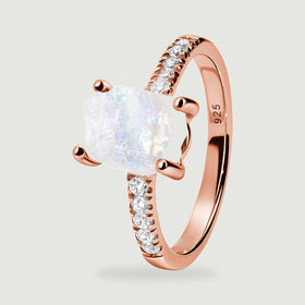 A rose gold vermeil ring is shown at an angle with an uneven pale moonstone, four prongs, and sparkling shoulder stones.