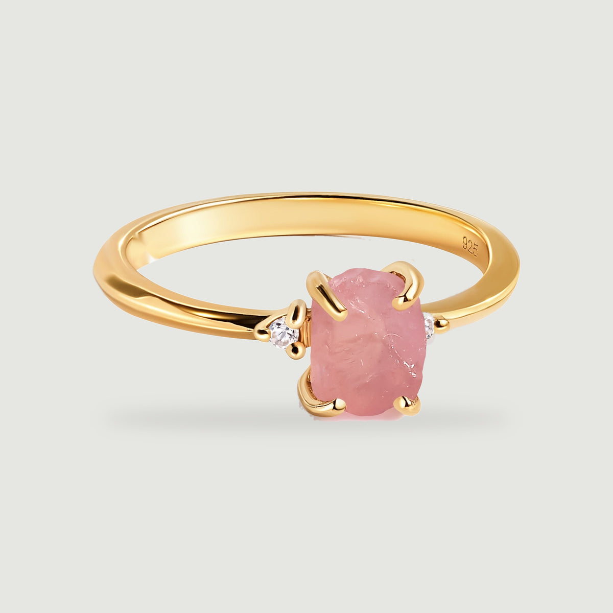 A yellow gold vermeil ring with a rough pink stone and two clear side stones rests on a light gray surface.