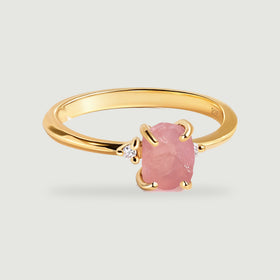 A yellow gold vermeil ring with a rough pink stone and two clear side stones rests on a light gray surface.
