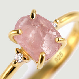A close view shows the uneven pink stone, four gold prongs and a clear round accent stone.