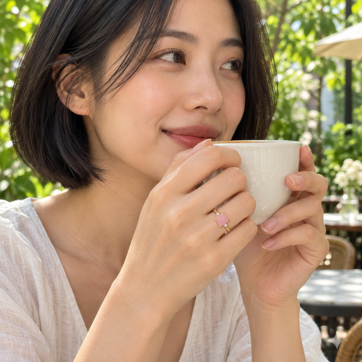 A woman holds a white ceramic cup with a pink stone ring on her hand at a leafy outdoor cafe.