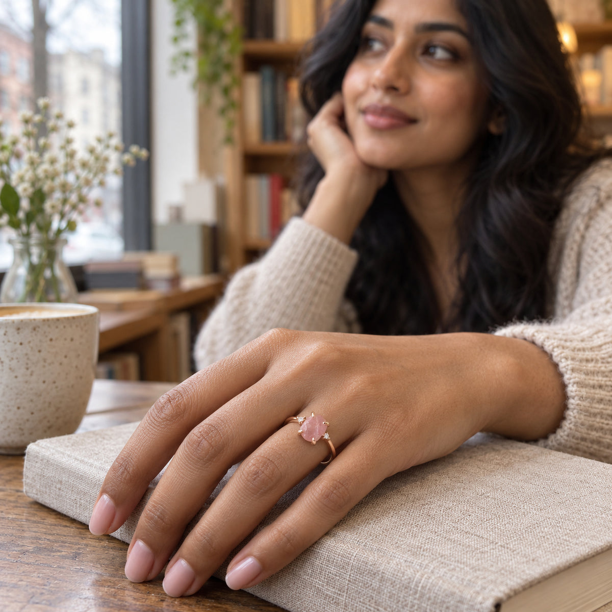 A rose gold ring with a rough pink stone is shown on a hand resting on a linen-covered book by a window.