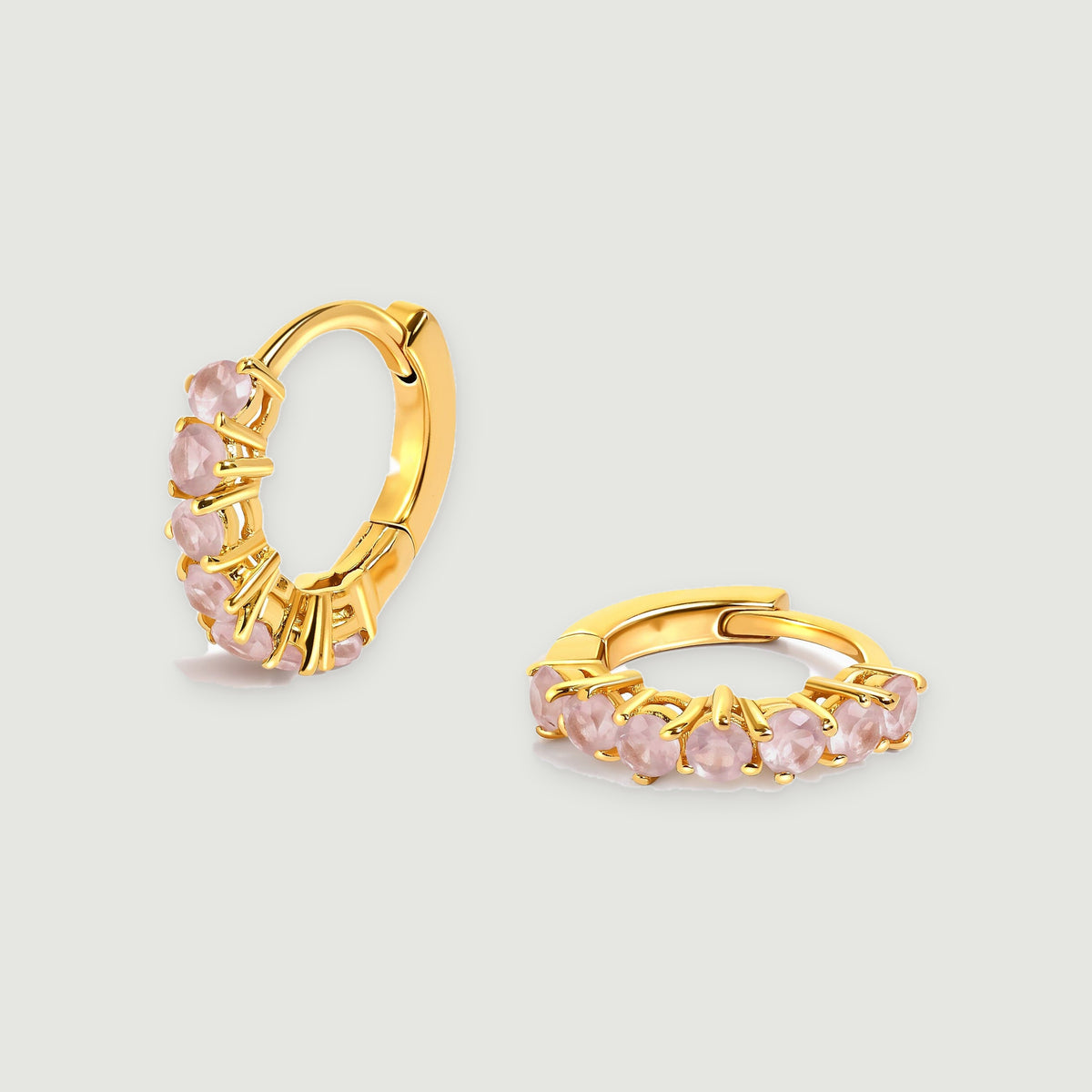 A pair of yellow-gold-tone hoops with pale pink round stones rests on a light gray background.