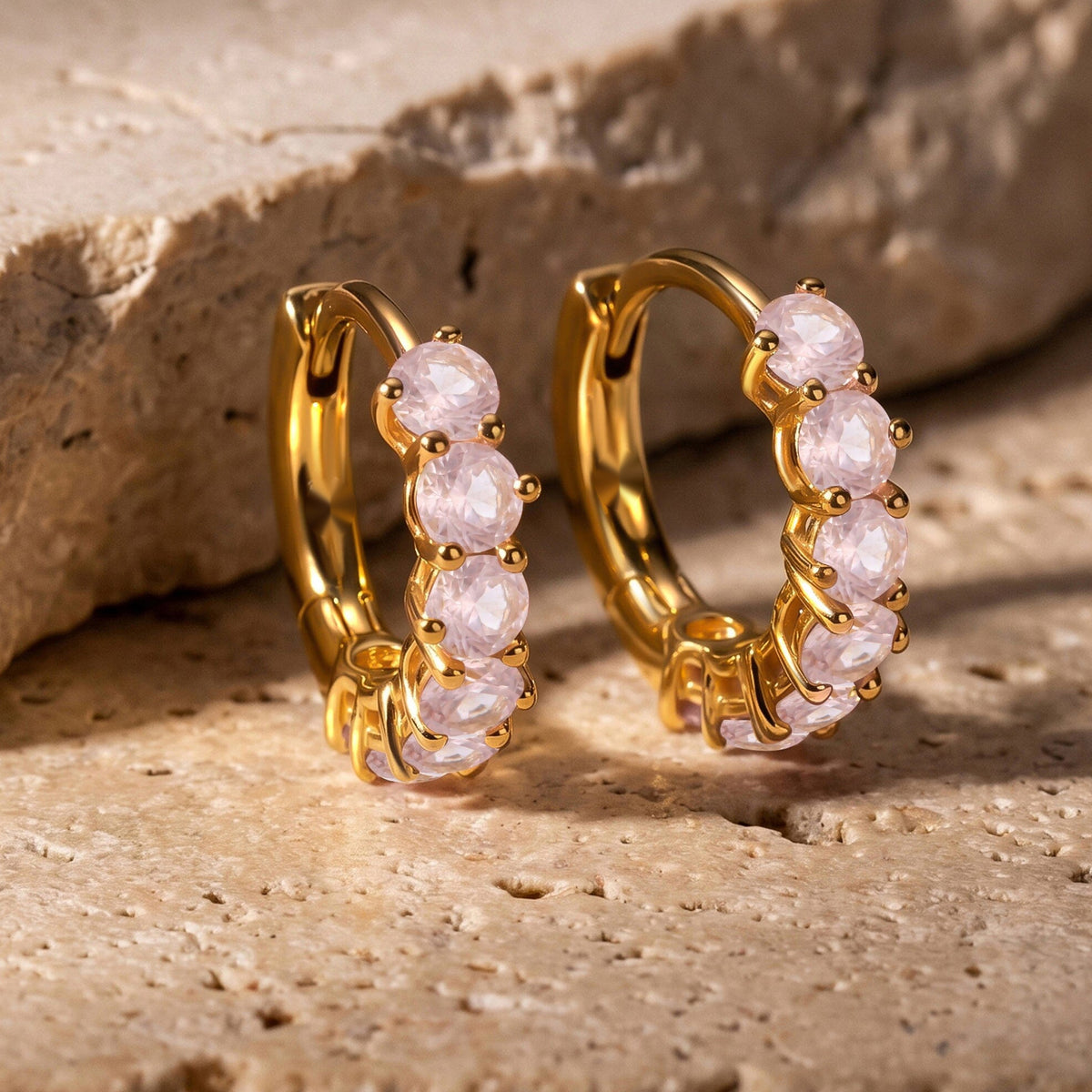 Two yellow-gold-tone rose quartz hoops stand beside a textured beige stone ledge.
