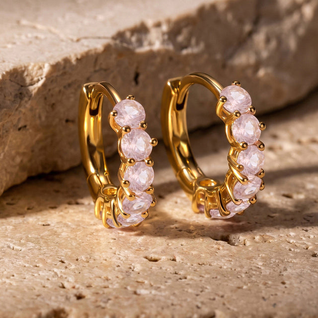 Two yellow-gold-tone rose quartz hoops stand beside a textured beige stone ledge.