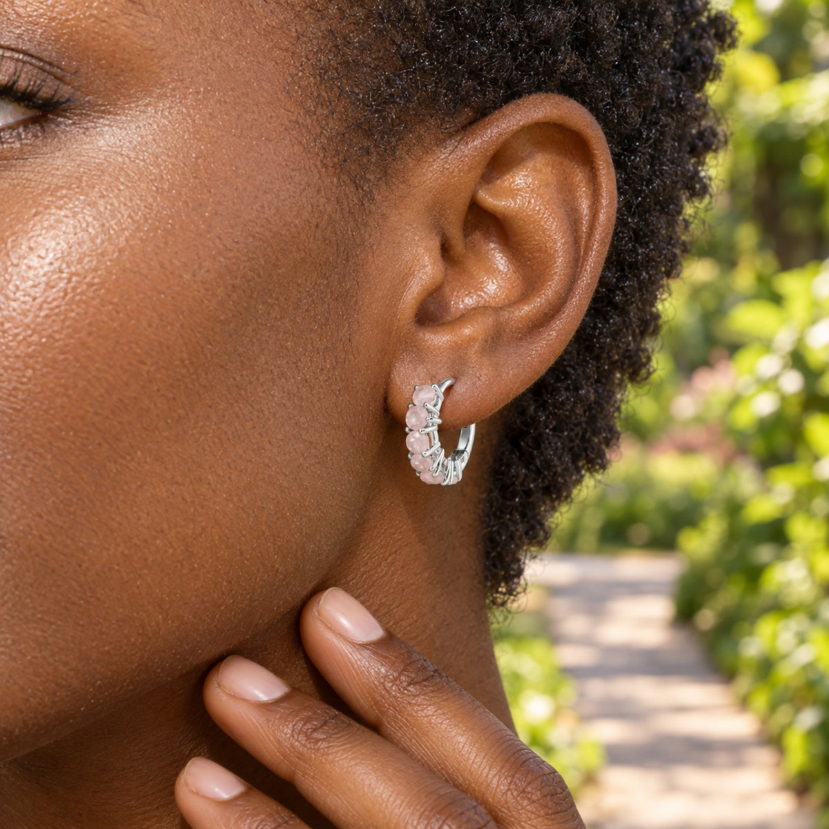 A woman with short coiled hair wears a silver-tone pale pink gemstone hoop beside a leafy garden path.