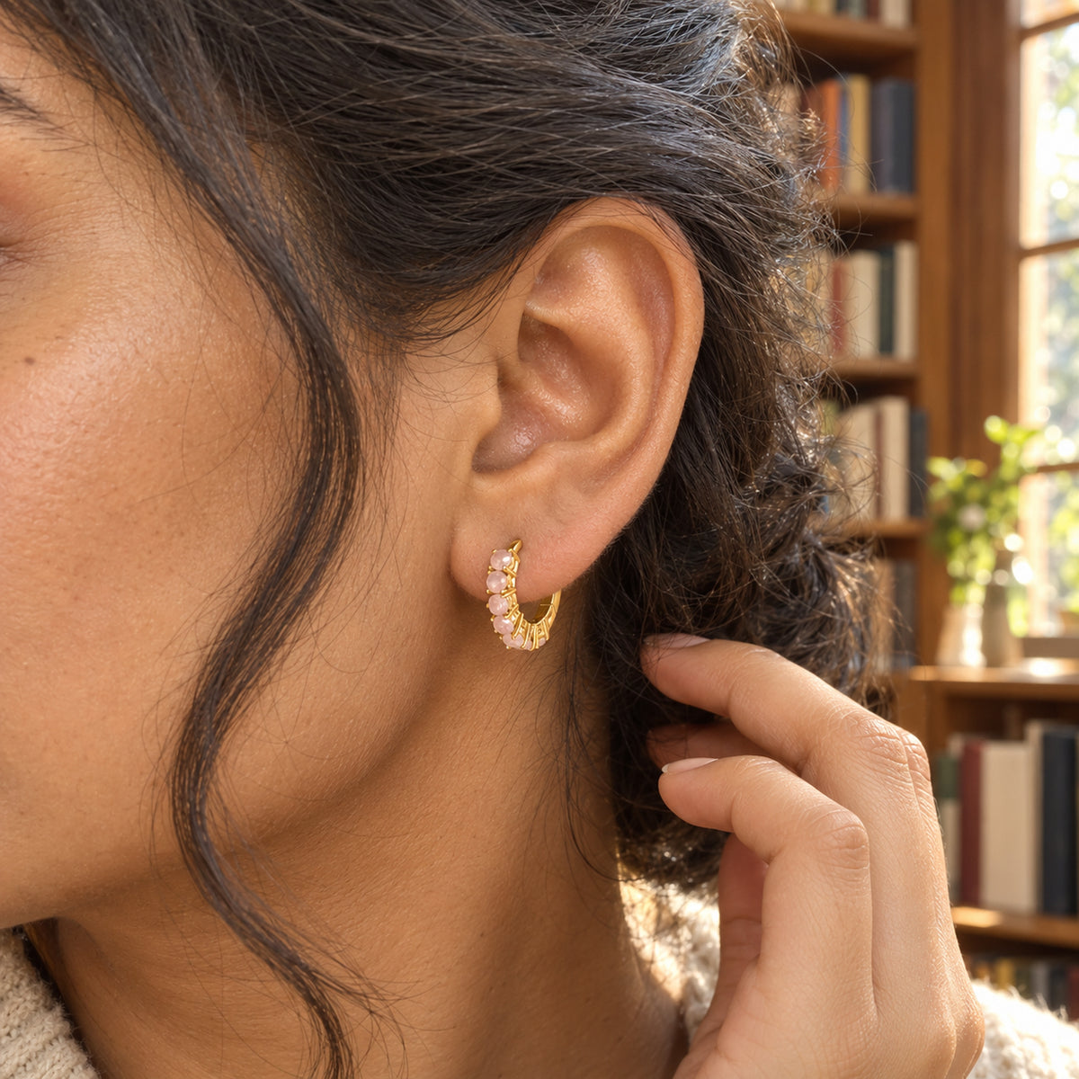 A woman tucks her dark hair behind an ear wearing a yellow-gold-tone rose quartz hoop inside a bookshop.