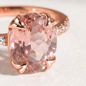Four rose gold prongs frame a pink faceted oval stone with clear accent stones along the curved band.