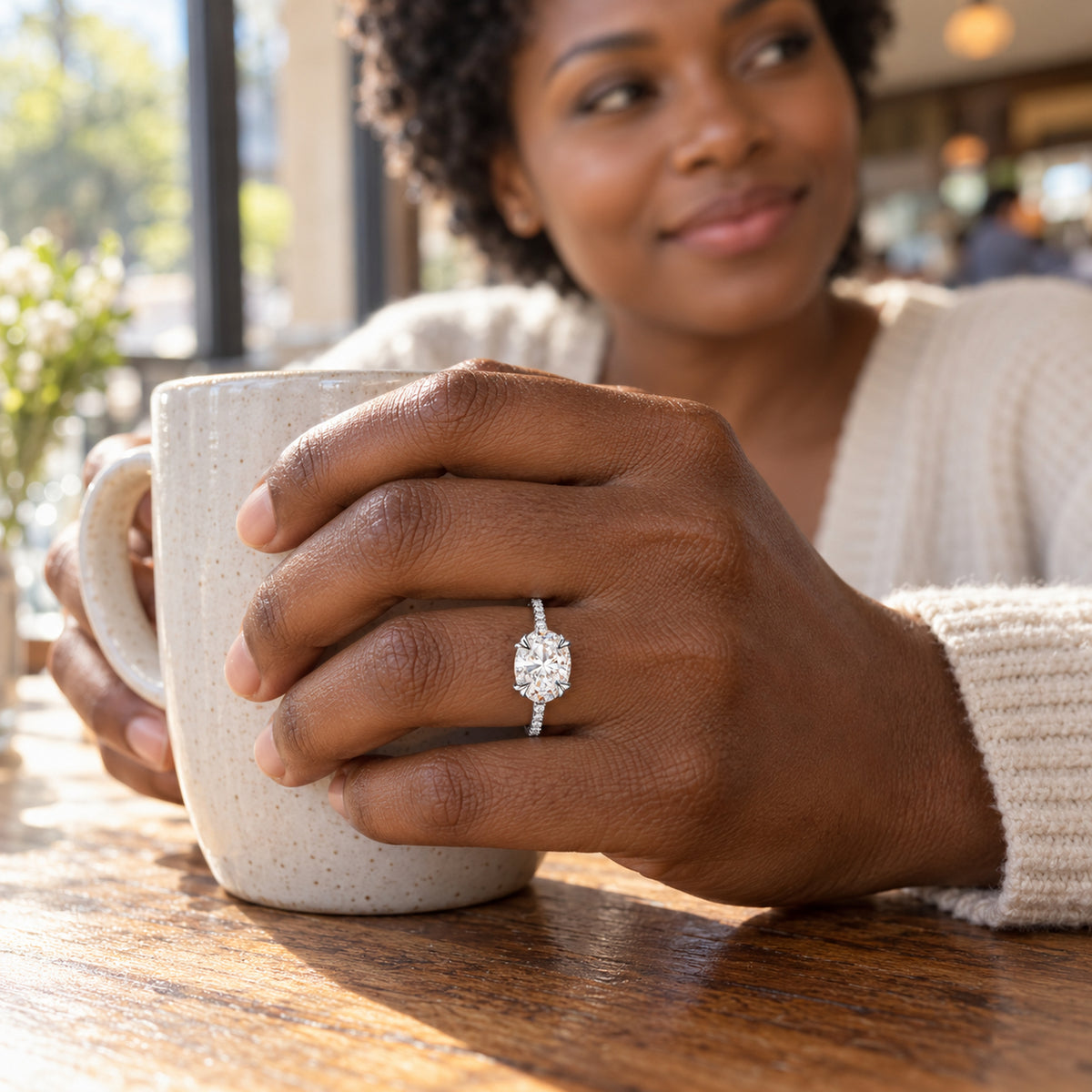 A sterling silver ring with a clear oval stone is worn on a hand holding a speckled ceramic mug at a cafe table.
