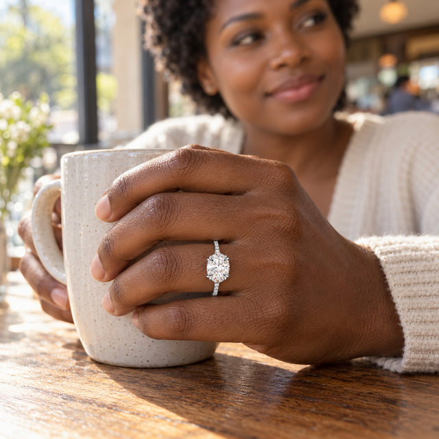A sterling silver ring with a clear oval stone is worn on a hand holding a speckled ceramic mug at a cafe table.