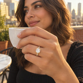 A yellow gold vermeil ring with an oval clear stone is worn on a hand holding a white cup on a terrace overlooking a city.