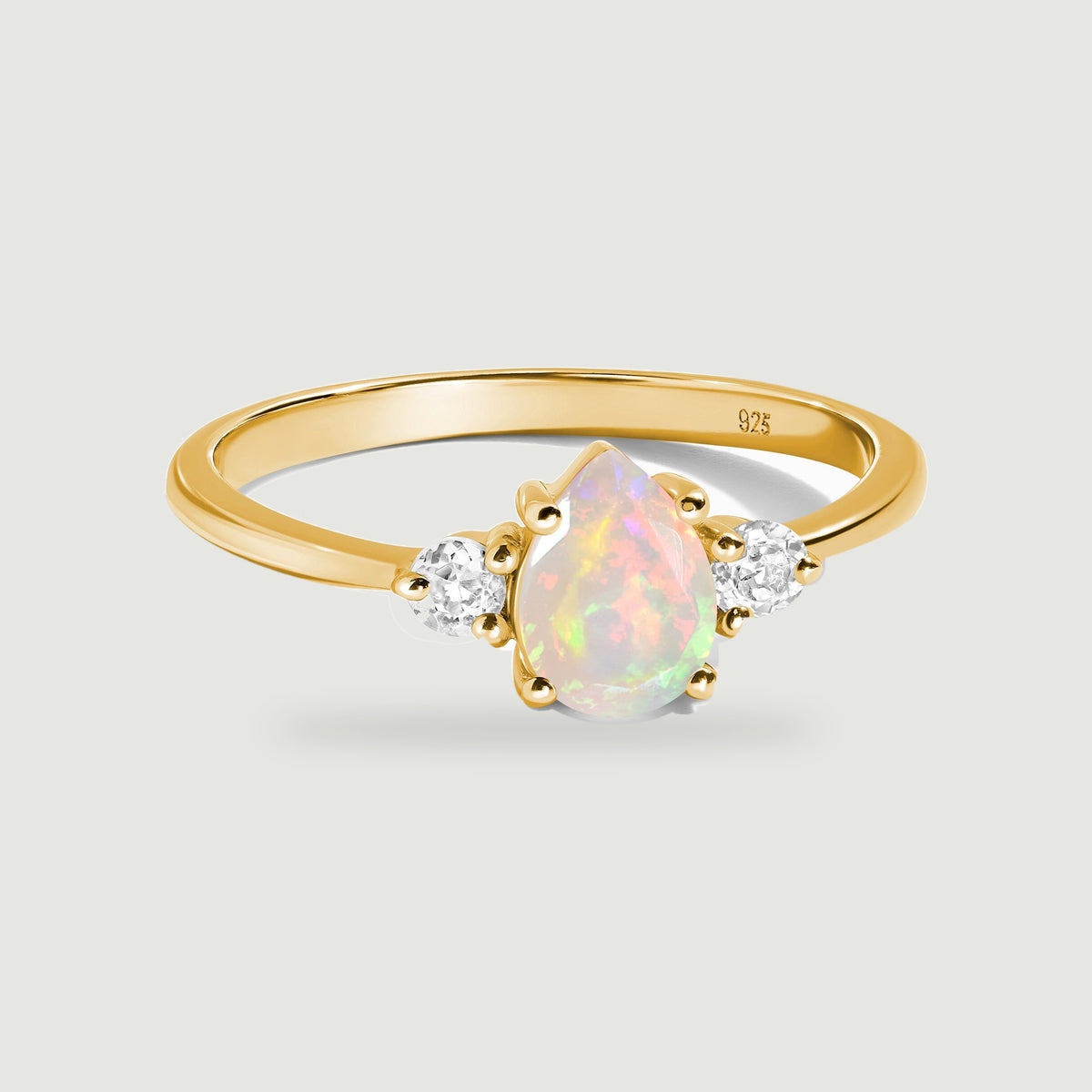 A yellow gold ring with a pear-shaped opal and two clear side stones rests on a light gray surface.