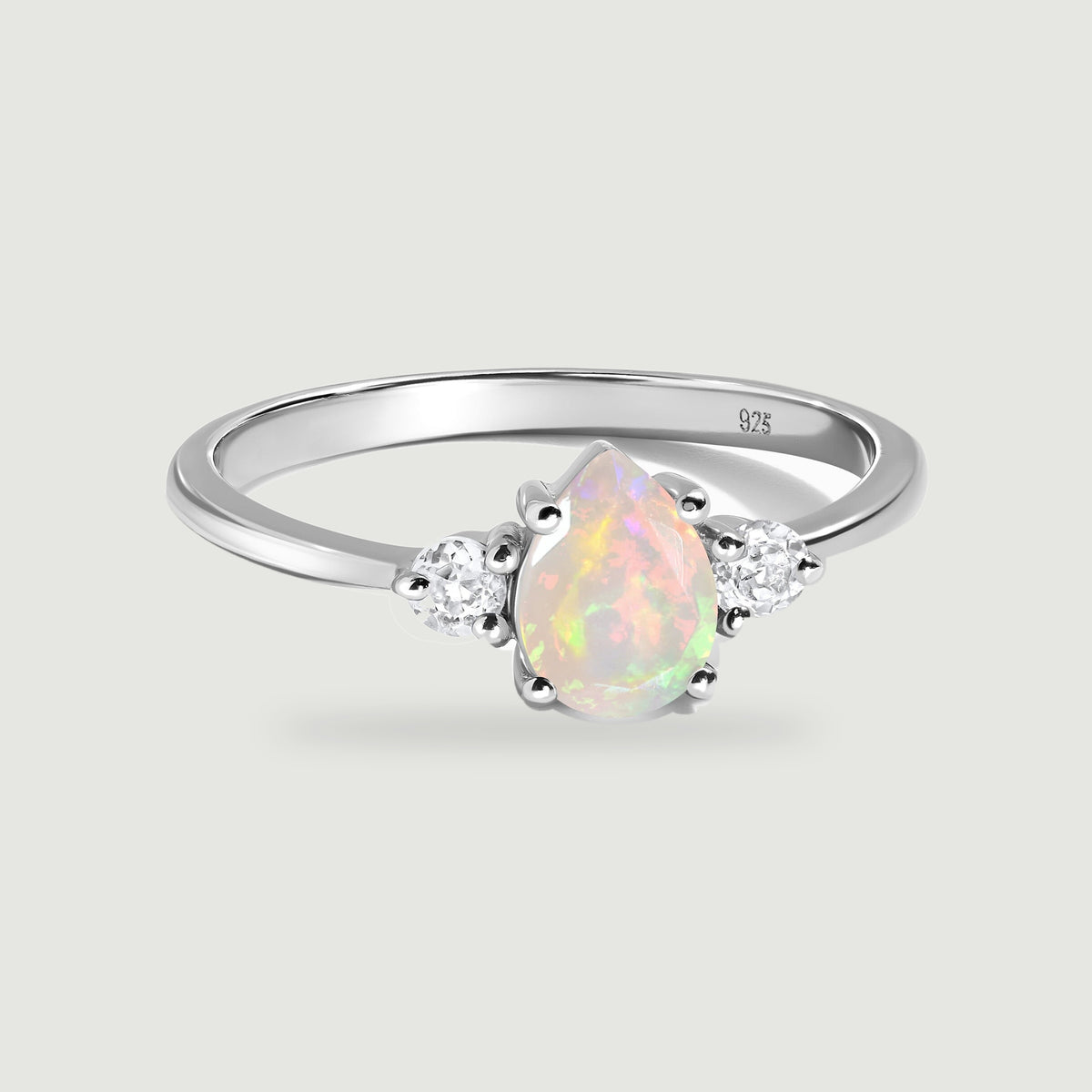 A silver ring displays a pear-shaped opal between two round clear stones on a pale gray background.