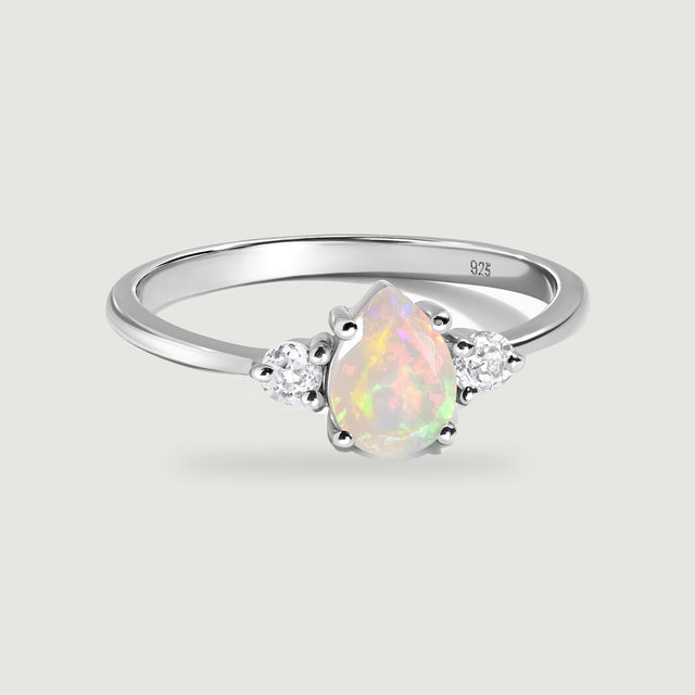 A silver ring displays a pear-shaped opal between two round clear stones on a pale gray background.
