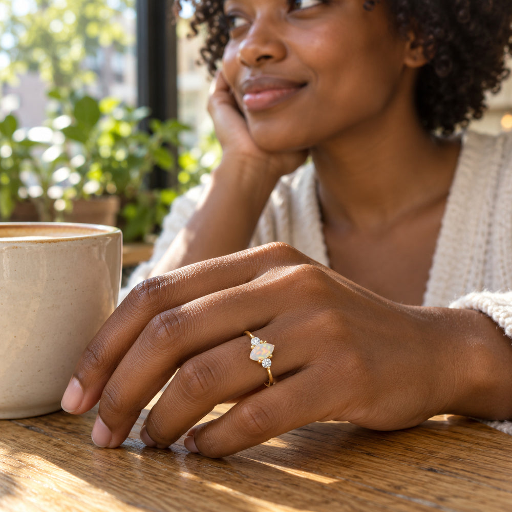A yellow gold opal ring is worn on a hand beside a ceramic cup at a wooden cafe table.