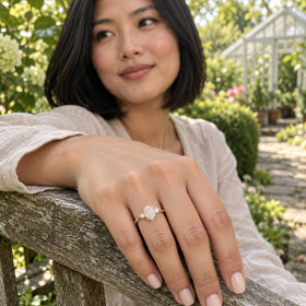A hand wearing the yellow gold opal ring rests on a weathered bench in a leafy garden.