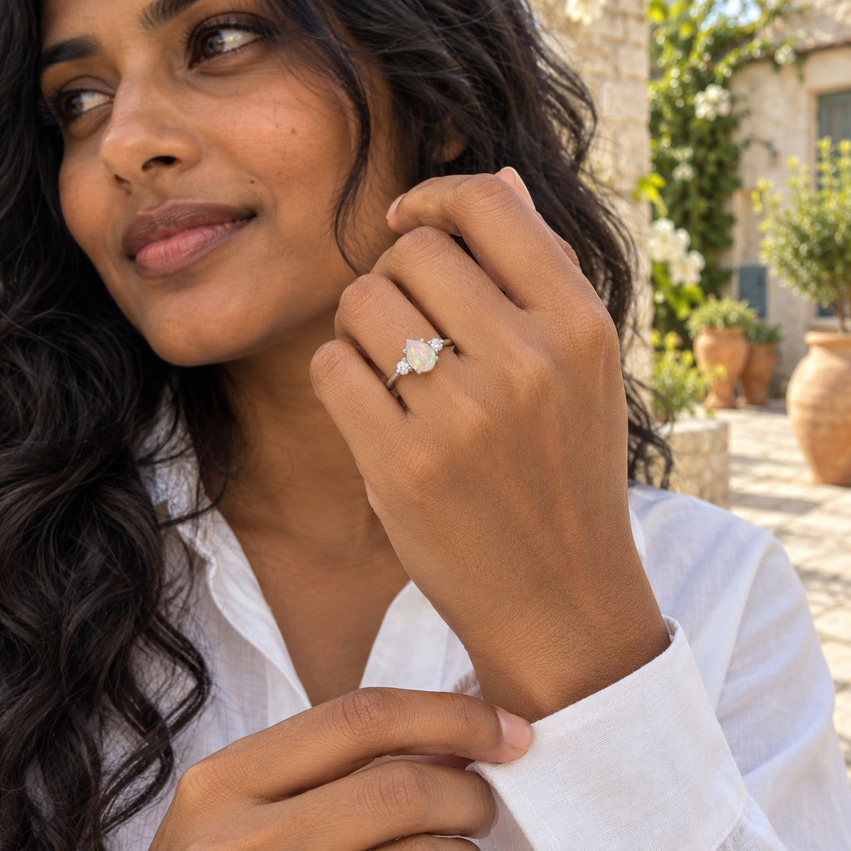 A silver opal ring is worn on a raised hand above a white shirt cuff in a stone courtyard.
