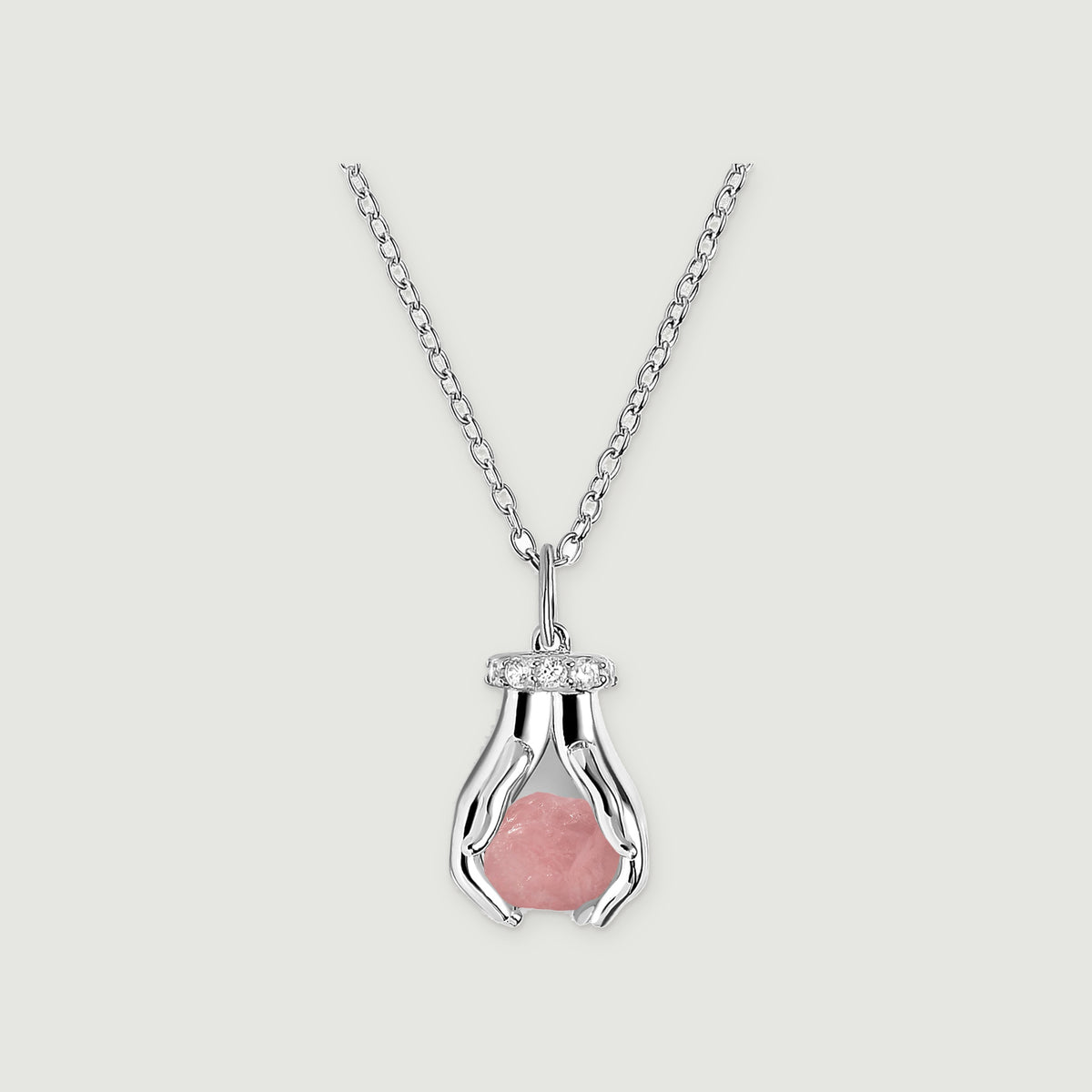 A sterling silver necklace with a raw pink quartz pendant framed by two sculpted hands and clear stones on a pale gray surface.