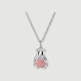A sterling silver necklace with a raw pink quartz pendant framed by two sculpted hands and clear stones on a pale gray surface.