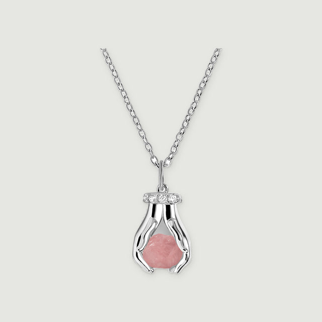 A sterling silver necklace with a raw pink quartz pendant framed by two sculpted hands and clear stones on a pale gray surface.