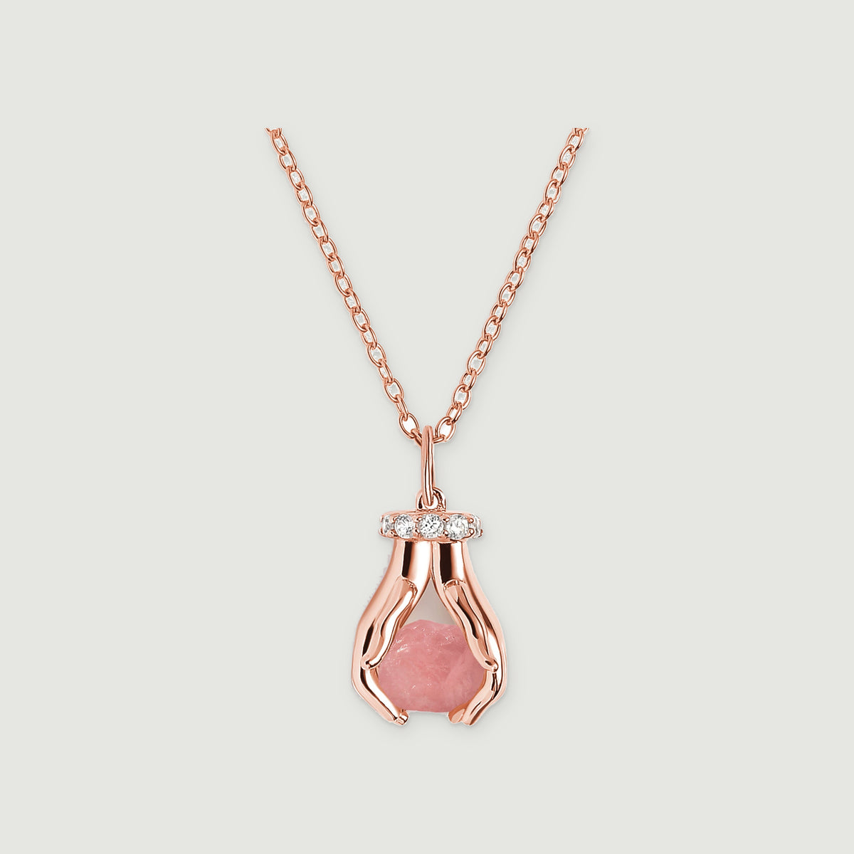 A rose gold vermeil necklace with a raw pink quartz pendant framed by two sculpted hands and clear stones on a pale gray surface.